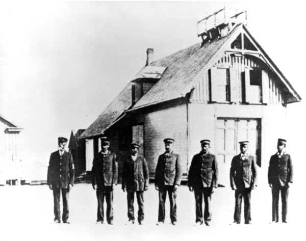 Photo of seven men in uniform posing in a row in front of the Pea Island Life-Saving Service Station.