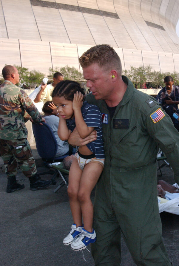Photo: A Coast Guard air crewmember carries a small child into a rescue helicopter. The crewmember is seen speaking with the child as it covers its ears from the noise. The crewmember has ear plugs.