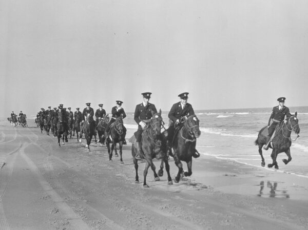 Black and white photograph of about 20 mounted beach patrol galloping along a beach.