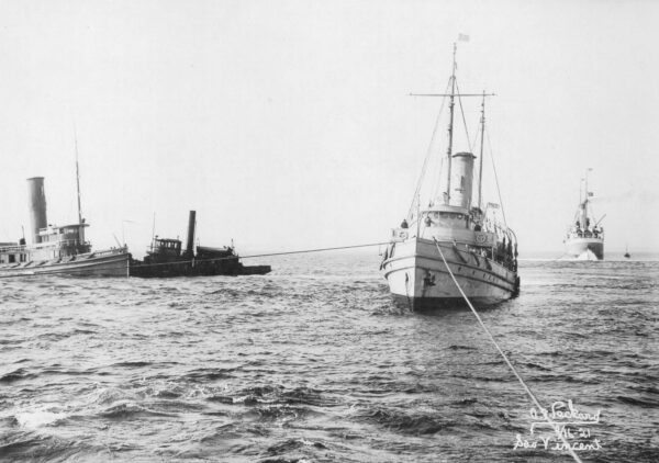 Photo of cutter Acushnet with several other ships. Heavy ropes connect it to a ship on its starboard side, as well another location, presumably from the vessel which the photograph was taken.