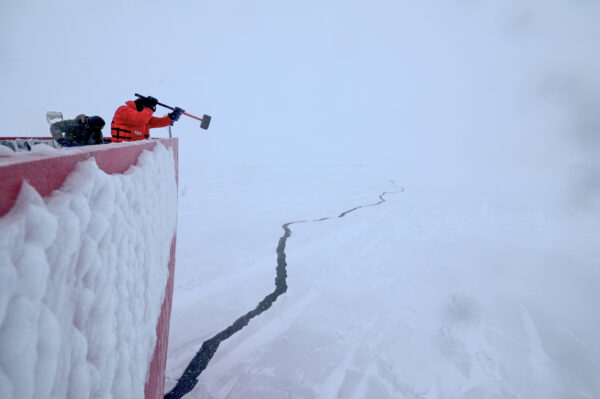 Photo: Crew works to remove heavy ice from the hull of Polar Star. In the background a large and dramatic crack forms in the ice leading off into the distance.