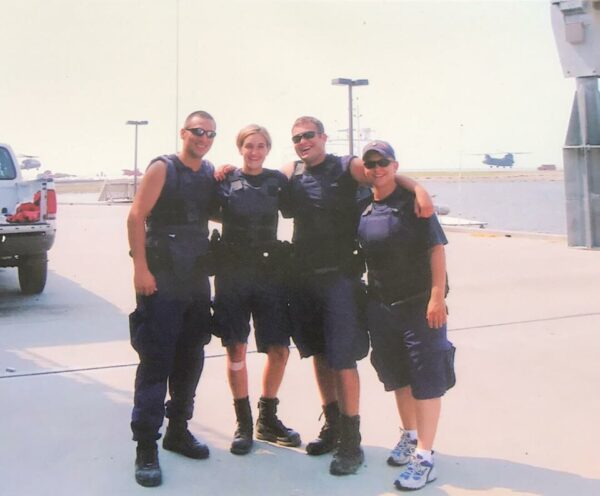 Photo: Two men and two women in blue uniforms posing with their arms around each other in a parking lot with a truck, part of a building, a helicopter and some lights in the background.