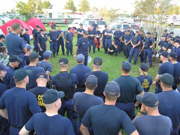 Photo: A Coast Guard lieutenant commander in a blue uniform stands on grass in middle of circle of more than 50 people in blue uniforms, surrounded by tents, cars and RVs.