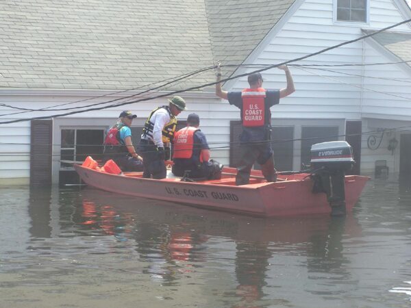 Photo: Four men in a boat next to a white house flooded almost to the roof. One man is standing holding power lines.
