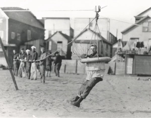 Black and white photo of a man in a breeches buoy on dry land.